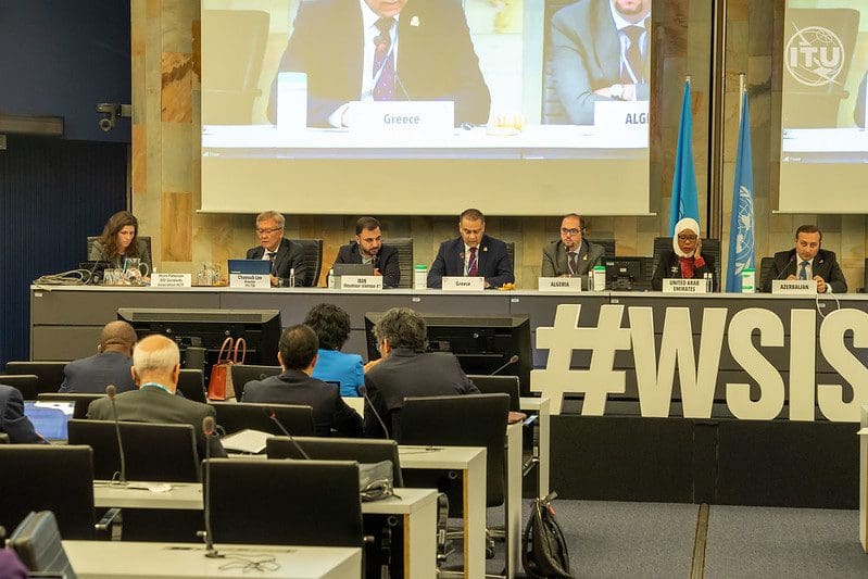Seven people sit as a panel in front of two light blue flags, one for the United Nations. The decorations in front of the table read #WSIS. The audience members sit at desks with microphones.