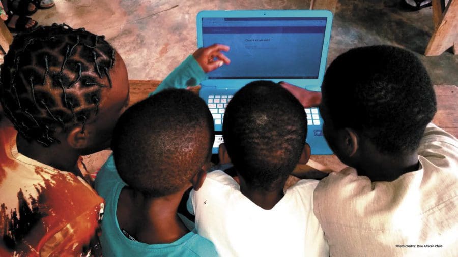 Four Black boys gather around a laptop on a wooden table. Text reads "Photo credits: African Child"