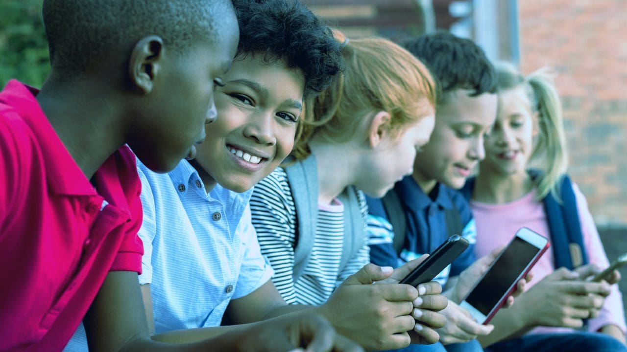 Children sitting together, smiling, playing on mobile devices.