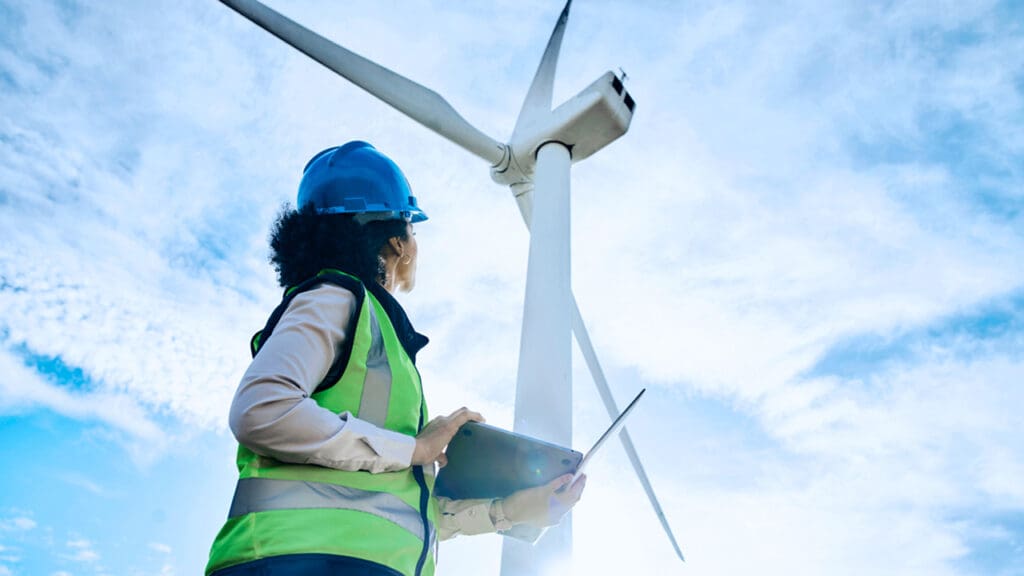 An engineer wearing a hardhat looks up at a wind turbine.